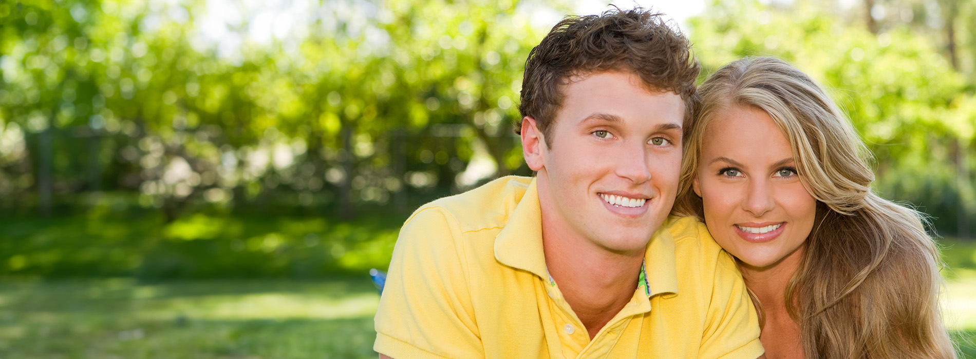 A young couple posing outdoors, with the man wearing a yellow shirt and the woman in a white dress, both smiling at the camera.