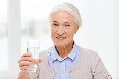 The image shows an elderly woman holding a glass of water with a smile on her face.