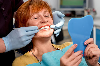 A woman with red hair is sitting in a dental chair, smiling at the camera while holding a blue dental impression model to her mouth, with a dental hygienist adjusting her teeth with a mirror.