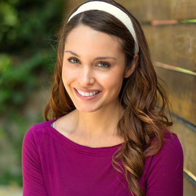 The image shows a young woman with long hair wearing a purple top, smiling at the camera, against a background featuring a wooden fence.