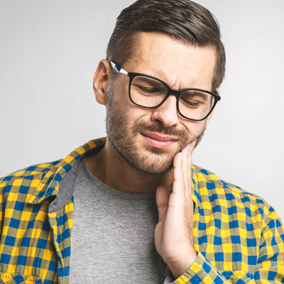 A man with glasses and a beard, wearing a yellow plaid shirt, appears distressed with his hand on his face, possibly indicating discomfort or pain.