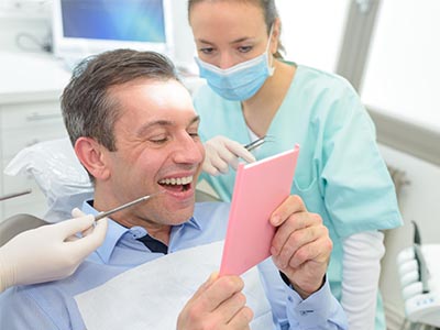 The image shows a man sitting in a dental chair, holding up a pink card with a surprised expression, while a dentist looks on, smiling.