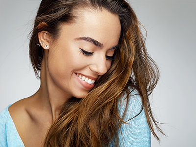 The image is a close-up portrait of a smiling woman with long brown hair, wearing a blue top, looking away from the camera with her gaze directed downwards.