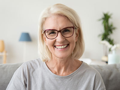 An elderly woman with blonde hair, glasses, and a light blue top, smiling at the camera.