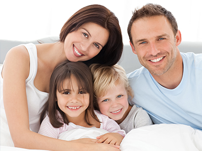 The image depicts a family of four people - an adult couple and their two children - smiling and posing together on a bed, with the father smiling directly at the camera while the mother and children look towards him.